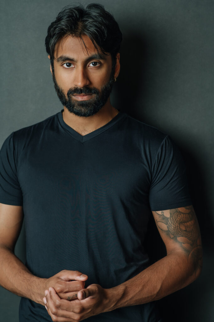 A man with beard and black shirt standing in front of a wall.